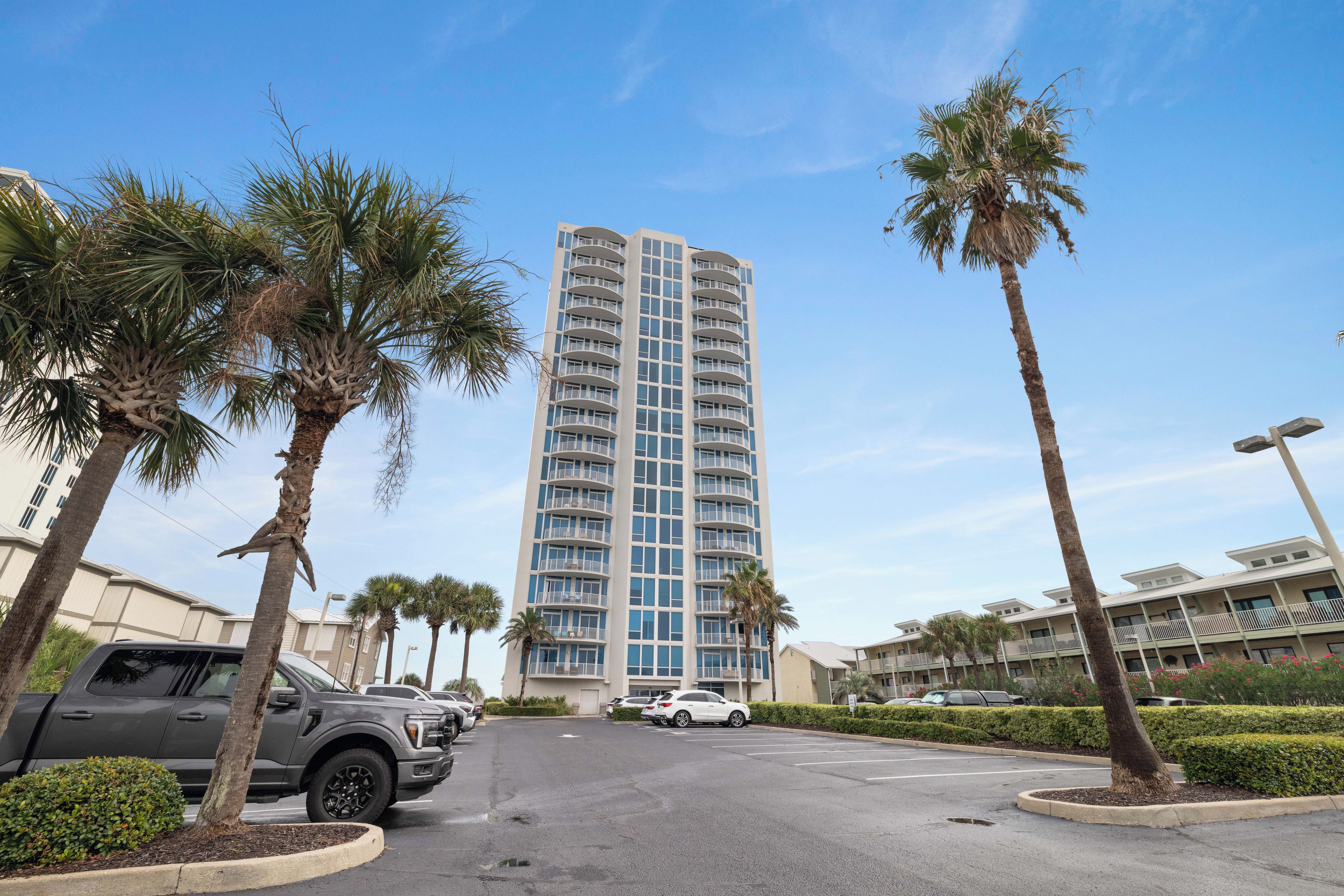 A multi-story hotel with a central courtyard, surrounded by palm trees and a parking area with cars.