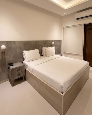 A modern bedroom with a large bed, bedside tables, a nightstand, and a wall-mounted air conditioning unit. - Hamsun Deluxe Double Room Clifton Karachi (Karachi)
