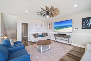Ein Wohnzimmer mit einem großen Flachbildfernseher, einem Deckenventilator und einem Gemälde zum Thema Strand.