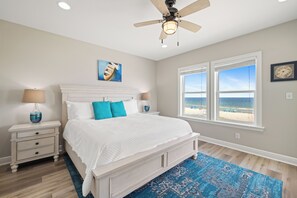 A bedroom with a bed, nightstands, a ceiling fan, a painting, a window with a beach view, and a patterned rug. - Bella Vista by Liquid Life (Gulf Shores)