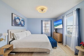 Une chambre à coucher comprenant un lit, une table de chevet, une commode, un téléviseur et une fenêtre avec des rideaux.