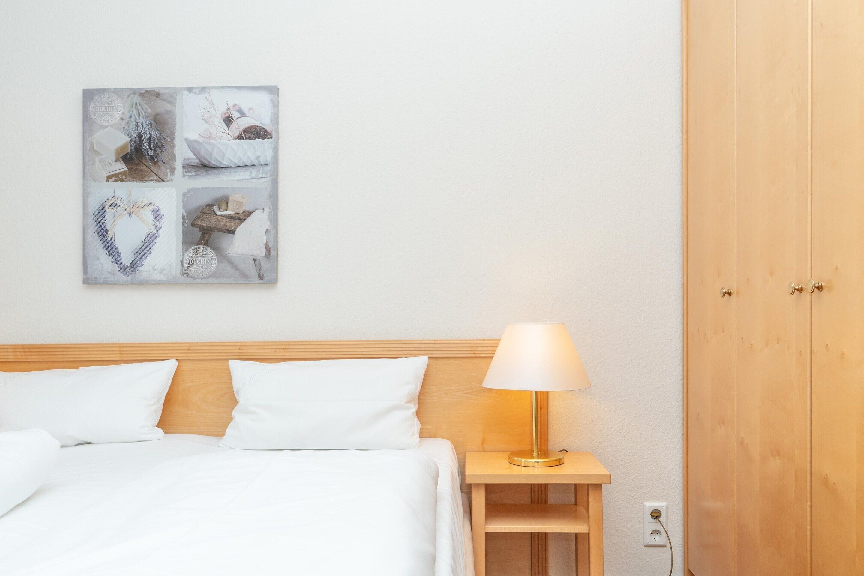 Ein ordentlich bezogenes Bett mit weißen Laken, einer Nachttischlampe und einem an der Wand hängenden Kunstwerk.