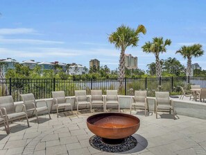 An outdoor seating area with a fire pit, chairs, and a view of a body of water and buildings in the background. - Coastal Blue | Summer Salt 24238 by Liquid Life (Orange Beach)