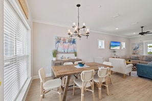 A dining area with a wooden table, wicker chairs, a chandelier, and a painting of palm trees. - Coastal Blue | Summer Salt 24238 by Liquid Life (Orange Beach)