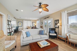 A spacious living room with a ceiling fan, a light fixture, a brown coffee table, and a wicker chair. - Gift By The Sea by Liquid Life (Fort Morgan)