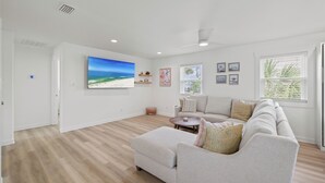 Een ruime woonkamer met een grote flatscreen-televisie, een beige bank, een salontafel en een raam met zonwering.
