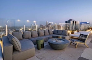 Eine Dachterrasse mit einer modernen Sofagarnitur, einem runden Couchtisch und Blick auf die Skyline der Stadt im Abendrot.