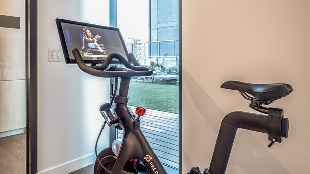 An indoor exercise bike with a screen displaying a woman, a wall-mounted control panel, and a view of a balcony with outdoor furniture. - Top Floor Downtown Lux Condo Rainey * Rooftop Pool (Austin)