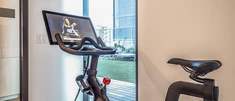 An indoor exercise bike with a screen displaying a woman, a wall-mounted control panel, and a view of a balcony with outdoor furniture.