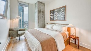 A bedroom with a large bed, a chair, a side table, a floor lamp, and a city view. - Rooftop Riverside Condo | *Rainey St*, Local Attractions, Rooftop Pool! (Austin)