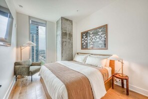 A bedroom with a large bed, a chair, a side table, a floor lamp, and a city view. - Rooftop Riverside Condo | *Rainey St*, Local Attractions, Rooftop Pool! (Austin)