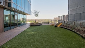 A modern building with large glass windows, a grassy courtyard, and a seating area with a yellow chair. - Rooftop Riverside Condo | *Rainey St*, Local Attractions, Rooftop Pool! (Austin)