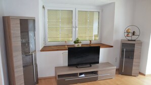 A modern living room with a flat-screen TV, wooden cabinets, and a window with blinds.