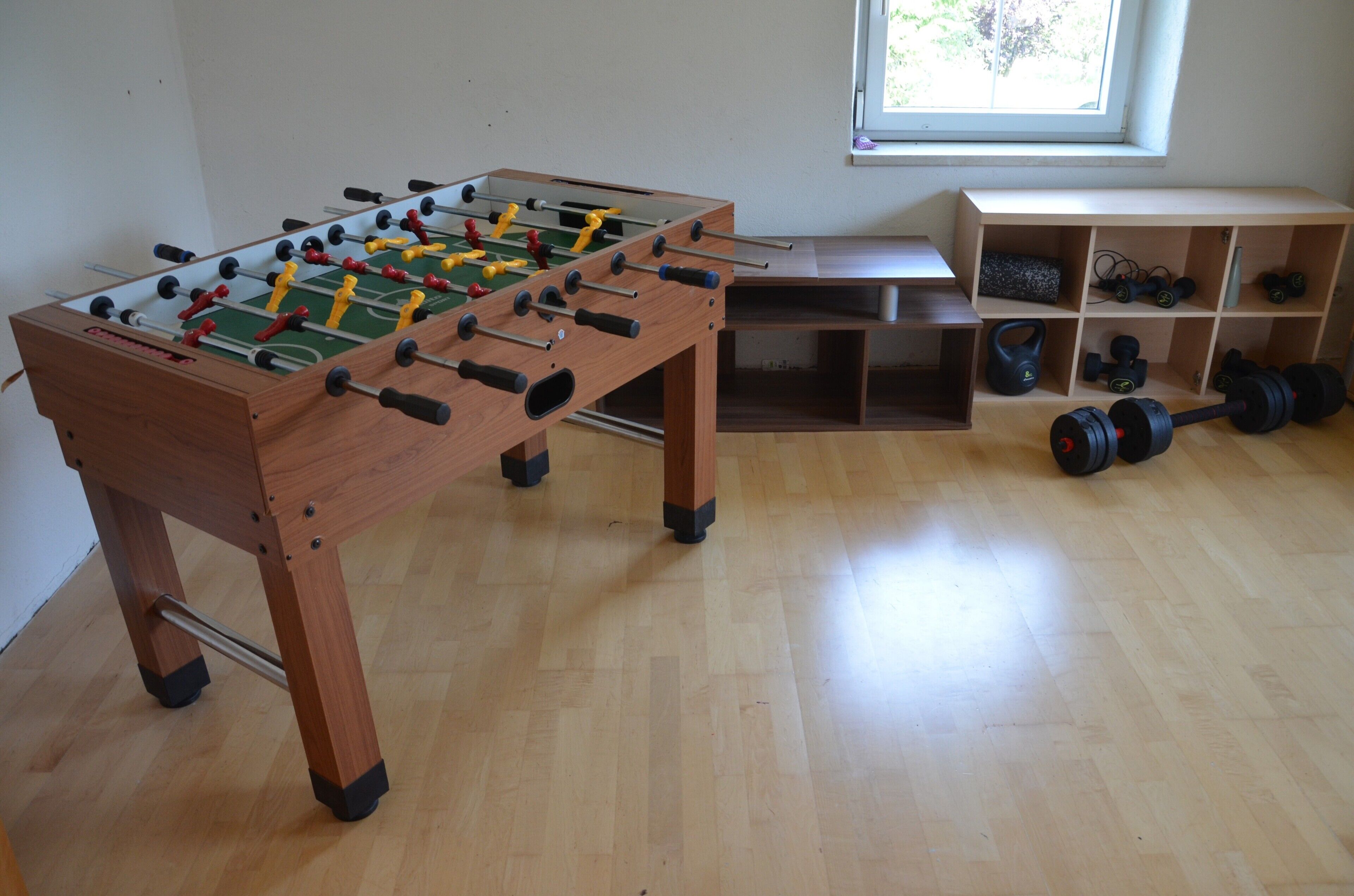 A wooden foosball table in a room with a window, shelves with weights, and a bench.