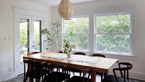 A dining room with a wooden table, six chairs, a hanging light fixture, and large windows with a view of greenery. - Luxe Venice Oasis | Abbot Kinney | Beach (Venice)