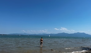 Eine Frau steht im flachen Wasser an einem Strand, im Hintergrund sind Berge zu sehen.