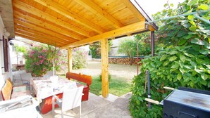 A covered patio with a table and chairs, a barbecue grill, and a garden area with plants and a wooden fence. - Holiday apartment Zoa (Pula)