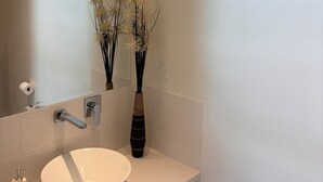 Une salle de bain avec un lavabo blanc, un miroir, un vase contenant des fleurs séchées et un distributeur de savon.
