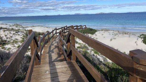 Une promenade en bois qui mène à une plage bordée de dunes de sable et d’où l’on a une vue dégagée sur l’océan.