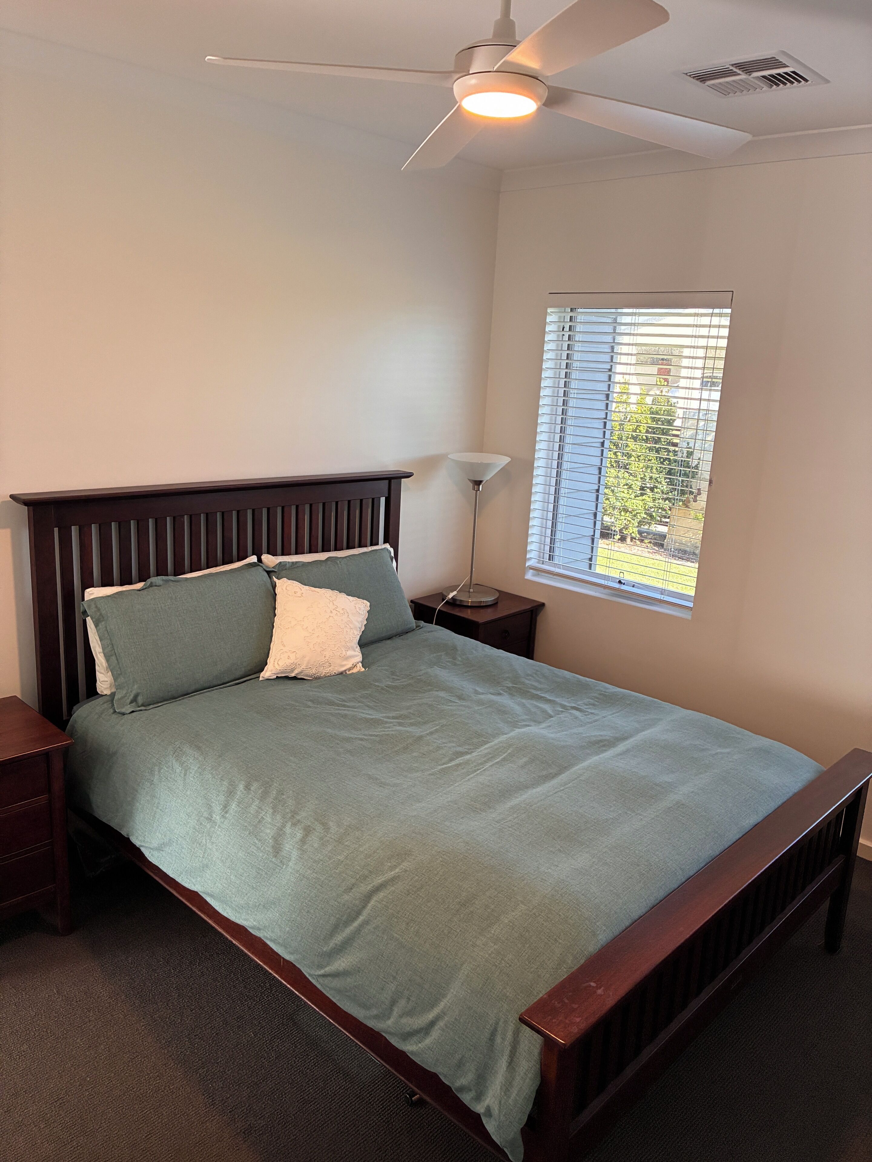 A bedroom with a bed, a nightstand, a lamp, a ceiling fan, and a window with blinds.