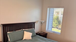 Une chambre à coucher comprenant un lit, une table de chevet, une lampe, un ventilateur de plafond et une fenêtre avec des stores.