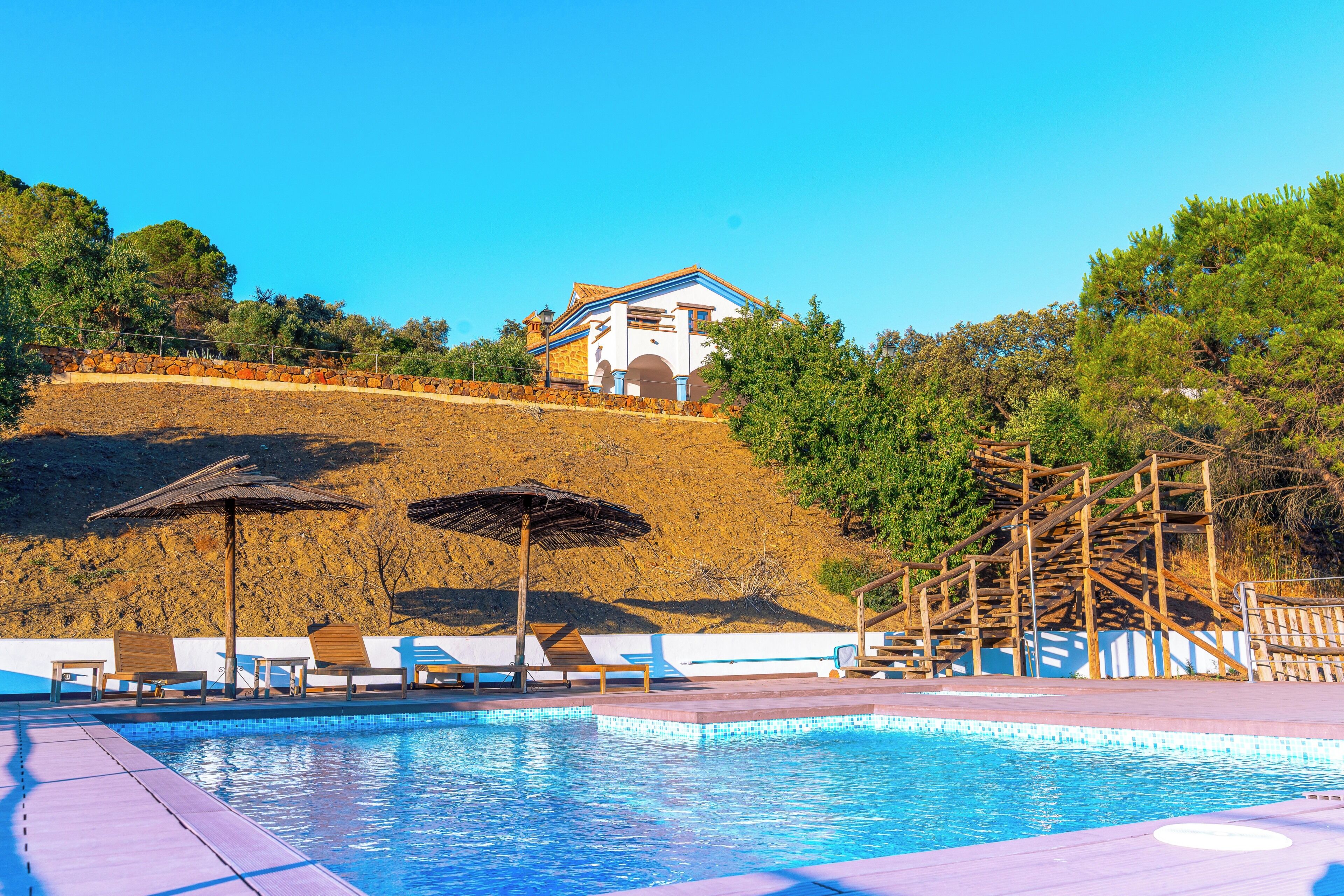 Une piscine avec des chaises longues et des parasols, un bâtiment avec un toit bleu et une colline parsemée d’arbres.