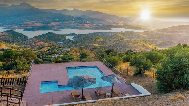 Piscina rectangular con dos sombrillas, una terraza de madera y vistas a montañas y un lago.