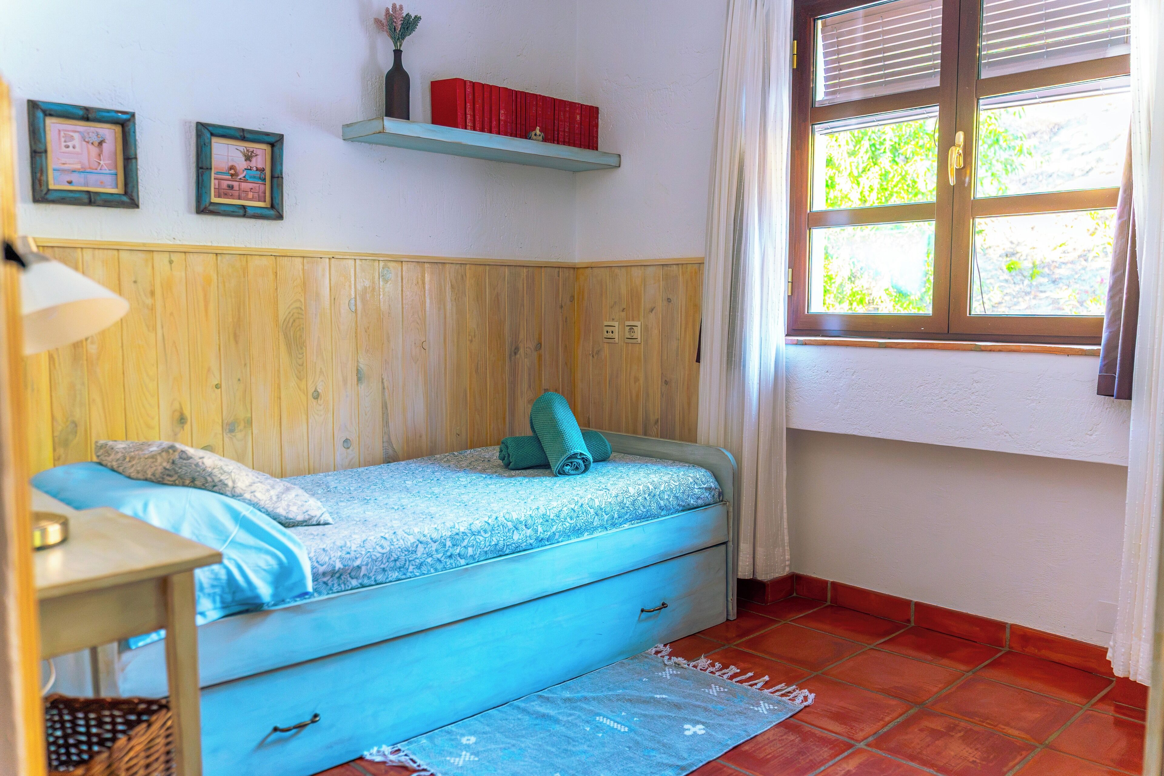 A room with a bed, a shelf with books, a window with blinds, and a wall with two framed pictures.