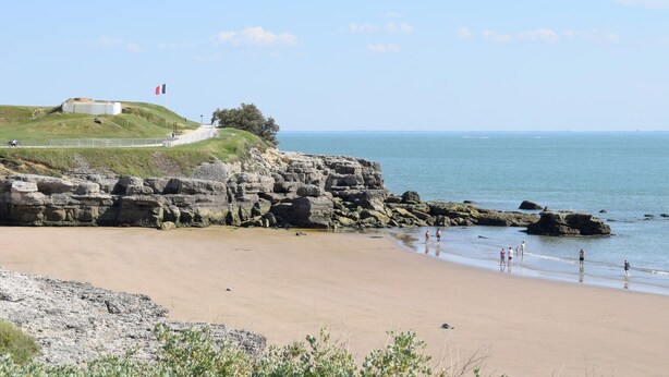 Un paysage côtier avec une plage de sable, des falaises rocheuses et un drapeau flottant au sommet d’une colline.