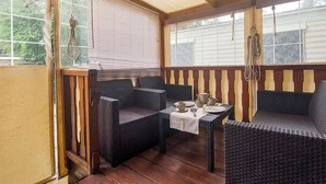 Une pièce avec un plafond en bois, une table recouverte d’une nappe blanche, deux chaises en osier et une fenêtre avec des stores.
