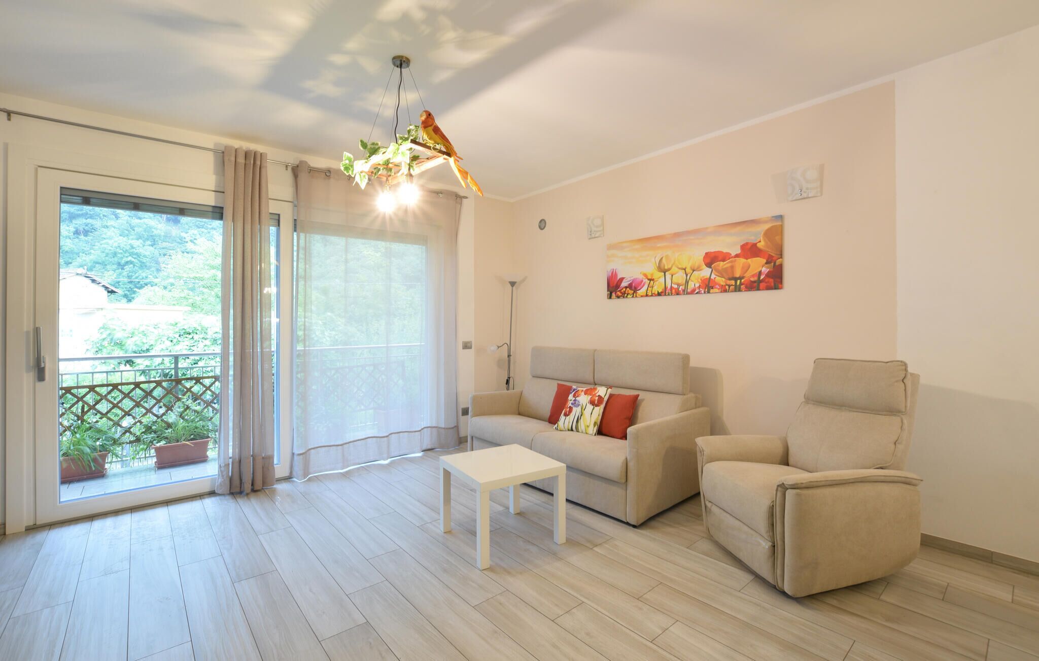 Ein Wohnzimmer mit Sofa, Sessel und Tisch. Ein Balkon mit Vorhängen und Blick ins Grüne.