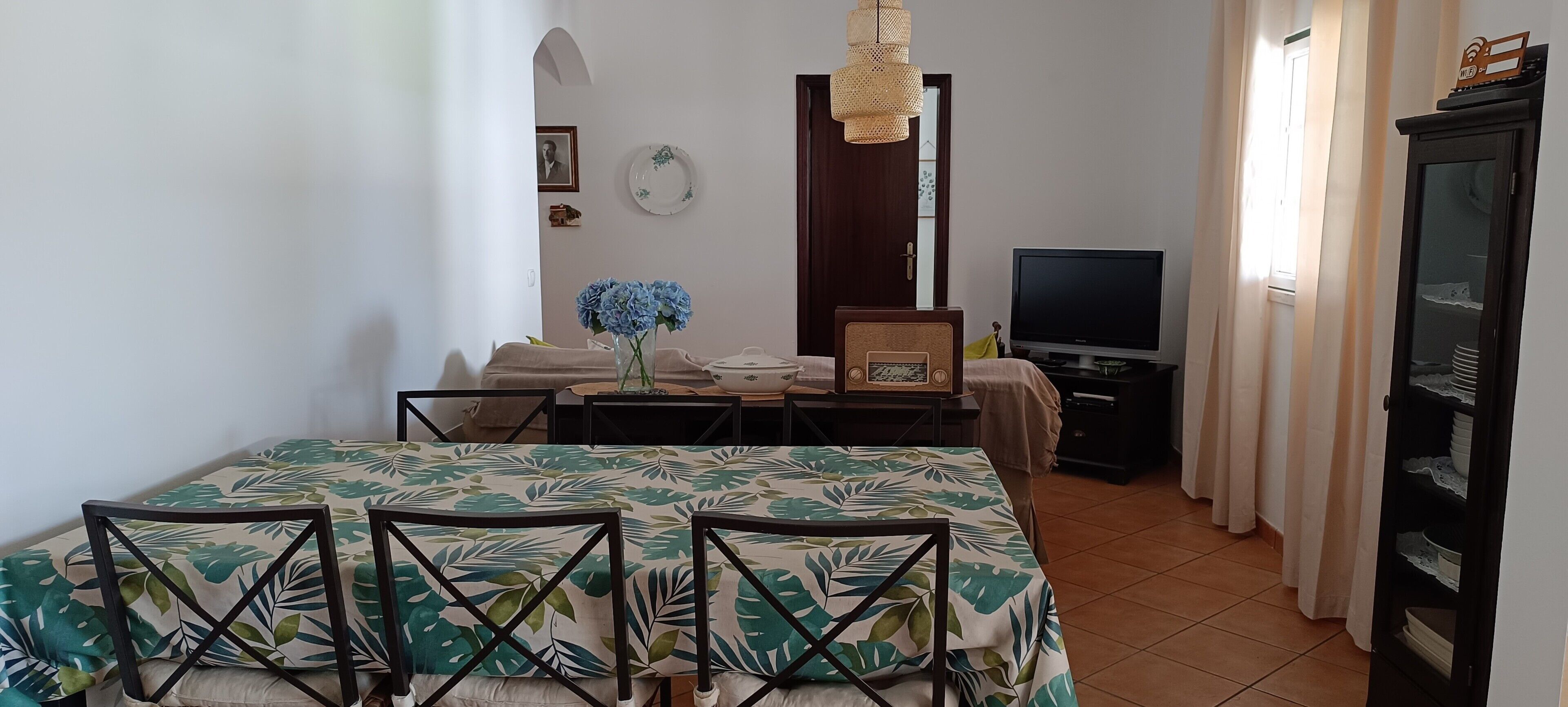 A room with a bed, a TV, a dining table with chairs, a vase with flowers, a clock, and a wardrobe.