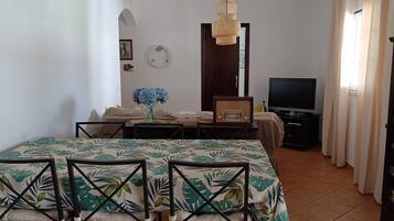 A room with a bed, a TV, a dining table with chairs, a vase with flowers, a clock, and a wardrobe.