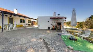 Un patio con suelo empedrado, zona de estar al aire libre con sillas y mesas, una parrilla y un porche cubierto.