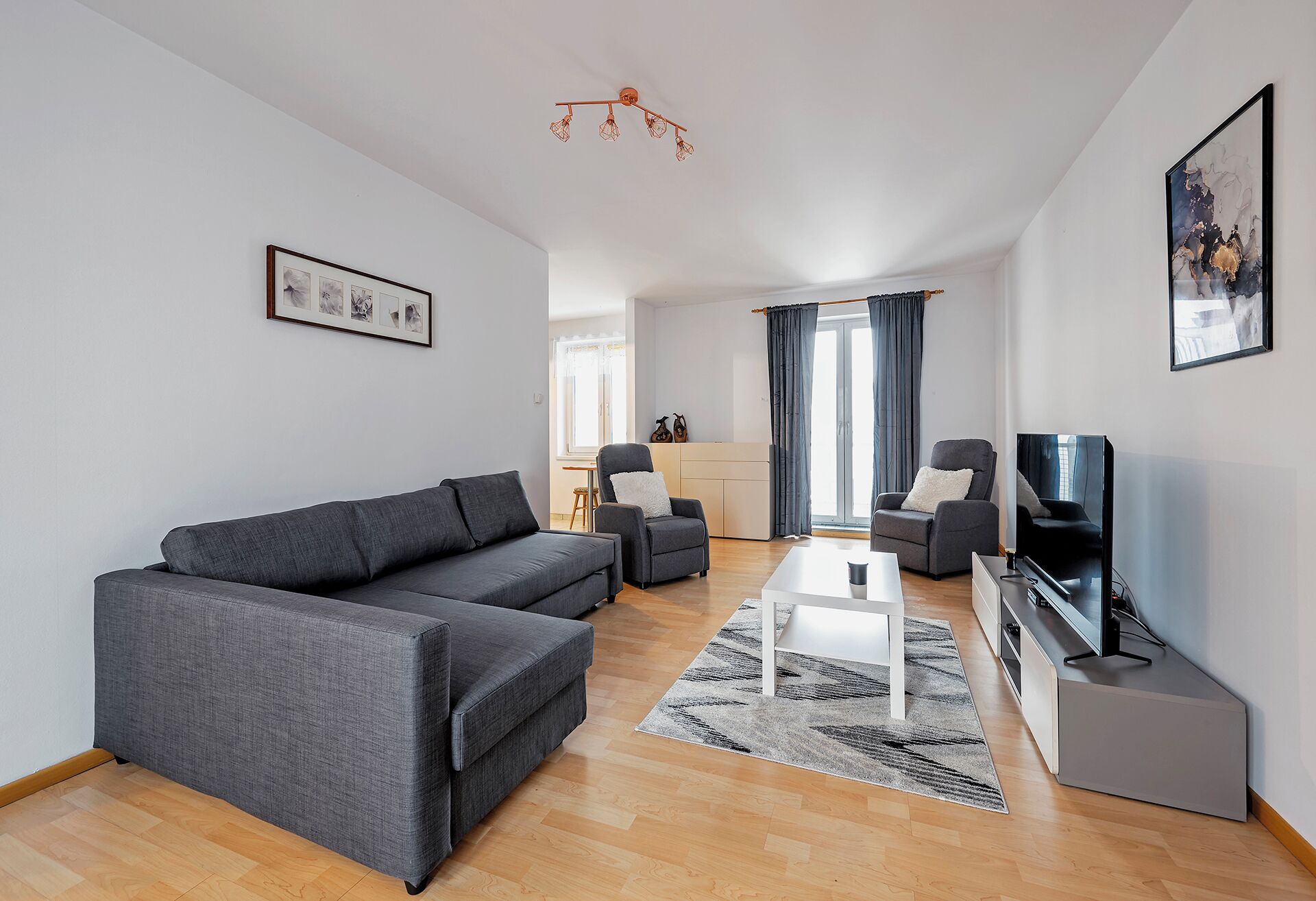 Un salon moderne avec un canapé gris, deux fauteuils, une table basse, un téléviseur et un tapis à motifs.