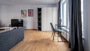 Uma sala de estar moderna com televisão de tela plana, sofá, mesa de jantar e uma cadeira. Há duas pinturas abstratas na parede e uma janela grande com cortinas.