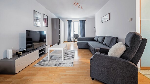 Uma sala de estar moderna com televisão de tela plana, sofá cinza, mesa de centro branca e uma janela grande com cortinas.