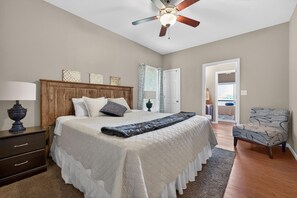 Quarto com cama, ventoinha no teto, mesa de cabeceira, cadeira e porta que dá para outro cômodo.