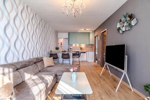 A modern living room with a sofa, a coffee table with a vase, a flat-screen TV on a stand, and a kitchen in the background. - Lelle Marine-hanna Pool Apartment (Balatonlelle)