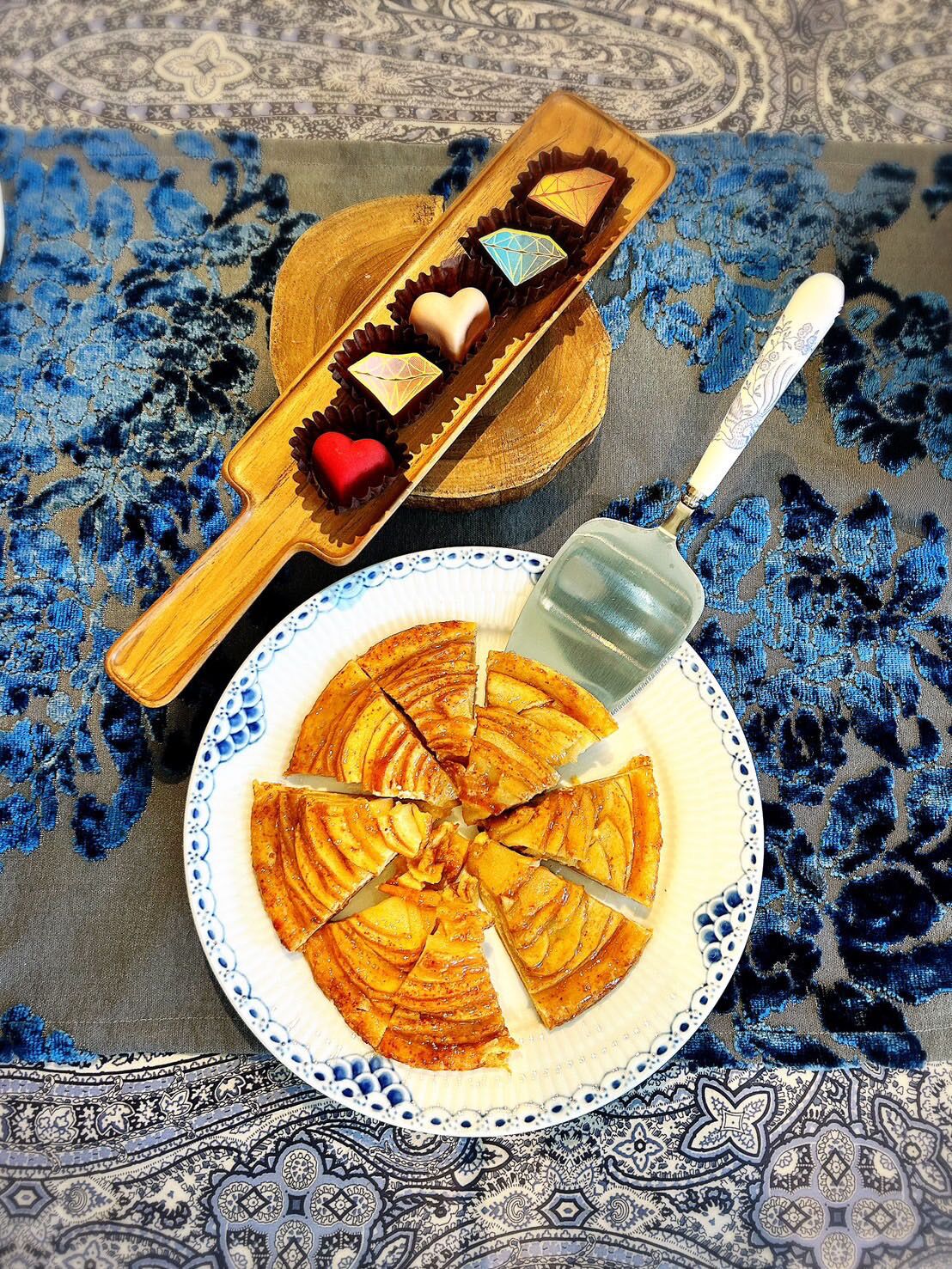 Plat de talls de poma, un ganivet, safata de fusta amb xocolates i una cullera decorativa sobre un mantell estampat.
