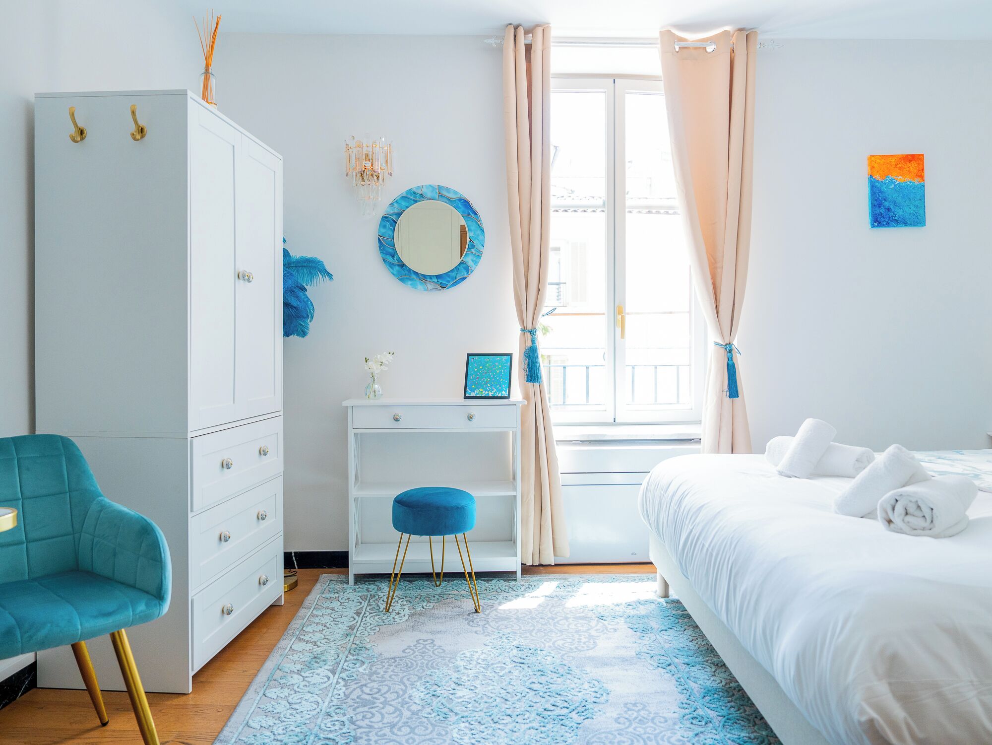 Une chambre à coucher comprenant un lit, une armoire, un bureau avec une chaise, une fenêtre avec des rideaux et un mur orné d’un miroir et d’œuvres d’art.