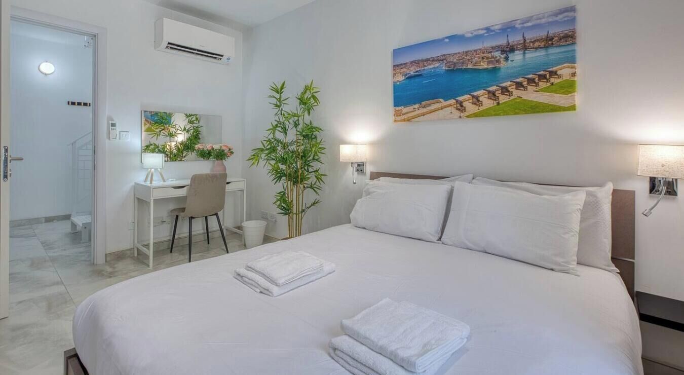 Una camera d'albergo con un letto, una scrivania, una sedia, una pianta e un quadro che raffigura una città costiera.