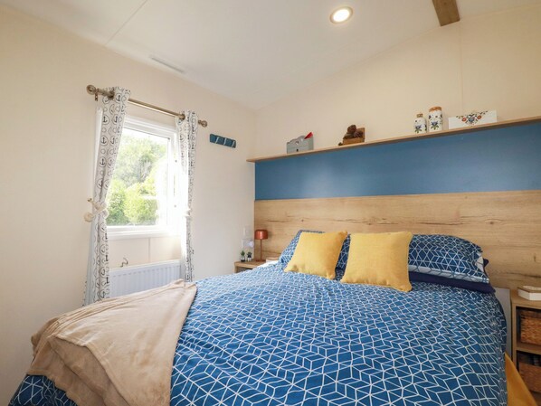 A bedroom with a bed, a window with curtains, a nightstand, and a shelf with decorative items. - Porthcurno 5 (Truro)