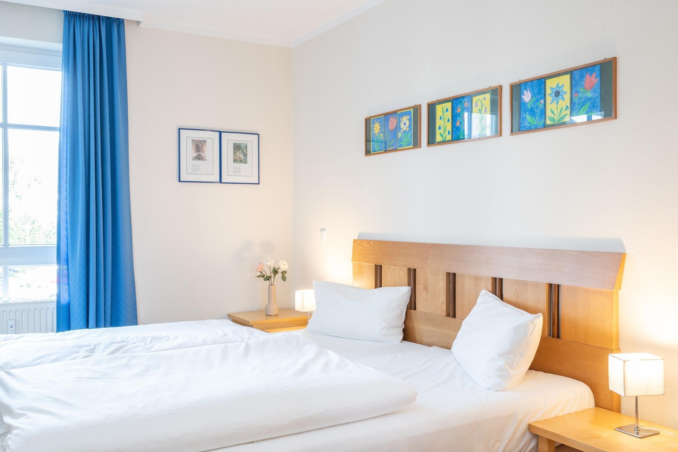 Quarto de hotel com cama de casal, mesa de cabeceira com luminária, janela com cortinas azuis e arte na parede.