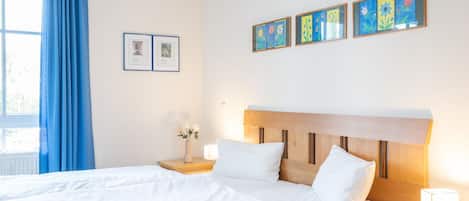 Quarto de hotel com cama de casal, mesa de cabeceira com luminária, janela com cortinas azuis e arte na parede.