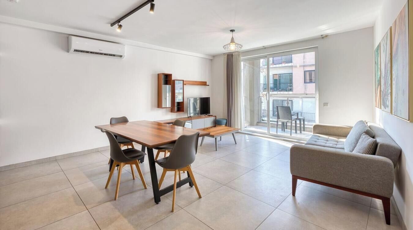 A modern living room with a sofa, a dining table with chairs, a TV, and a balcony with a view.