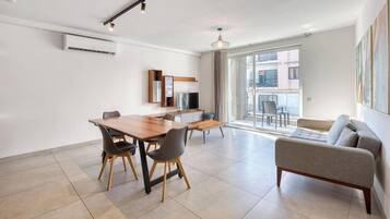 A modern living room with a sofa, a dining table with chairs, a TV, and a balcony with a view.