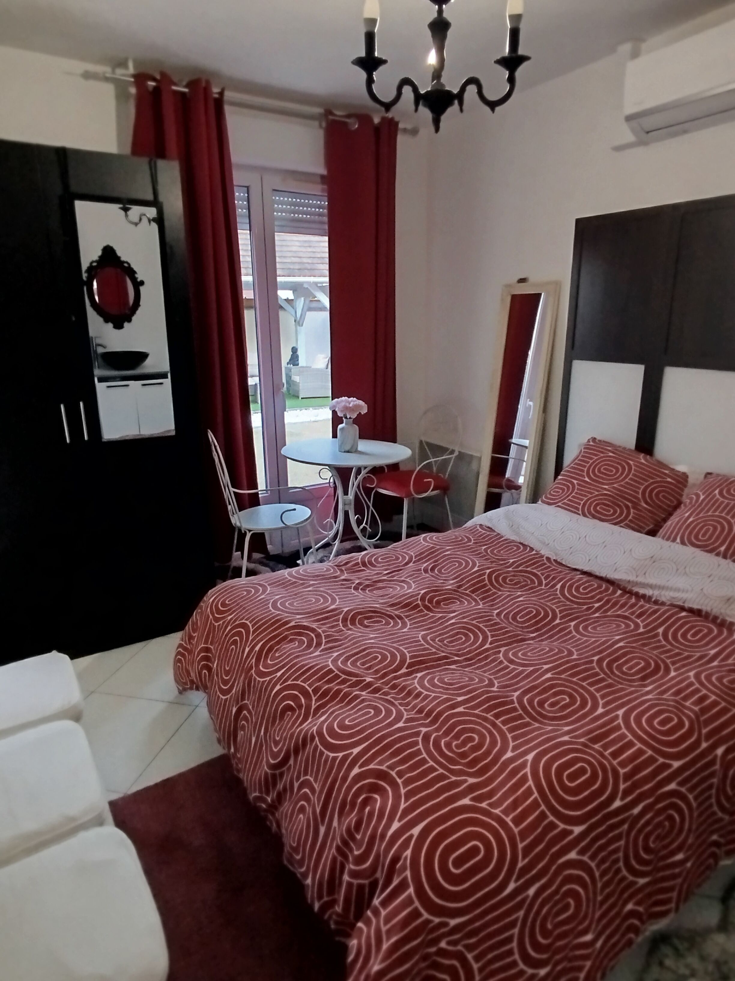 Ein Schlafzimmer mit einem Bett, einem kleinen Tisch, einem Sessel, einem Spiegel und einem Fenster mit Vorhängen.