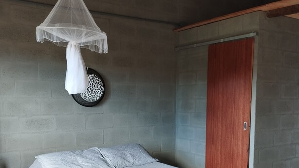 Un dormitorio con una cama, mosquitera colgante, un adorno redondo, una puerta de madera y una pared de concreto.