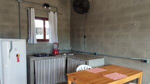 Una cocina compacta con refrigerador, estufa y lavamanos, una mesa de madera con sillas y un ventilador en la pared.